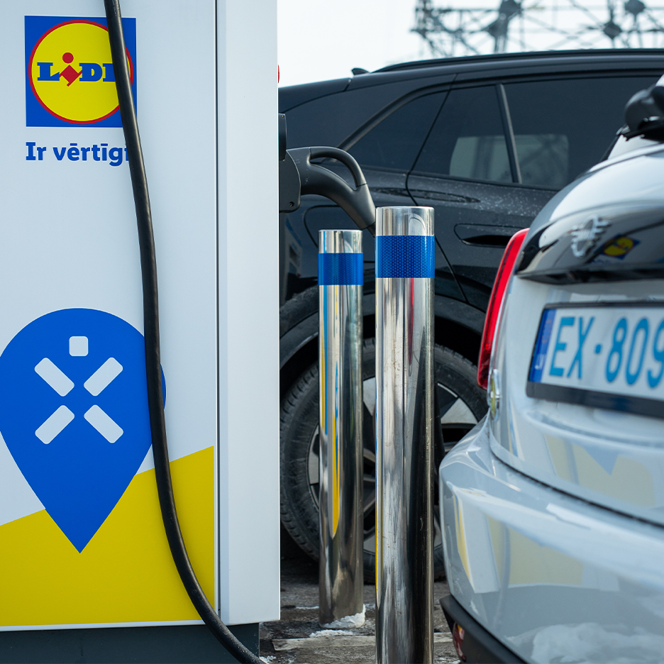 Elektromobiļi uzlādējas stacijā, blakus kurai redzams veikala logotips.