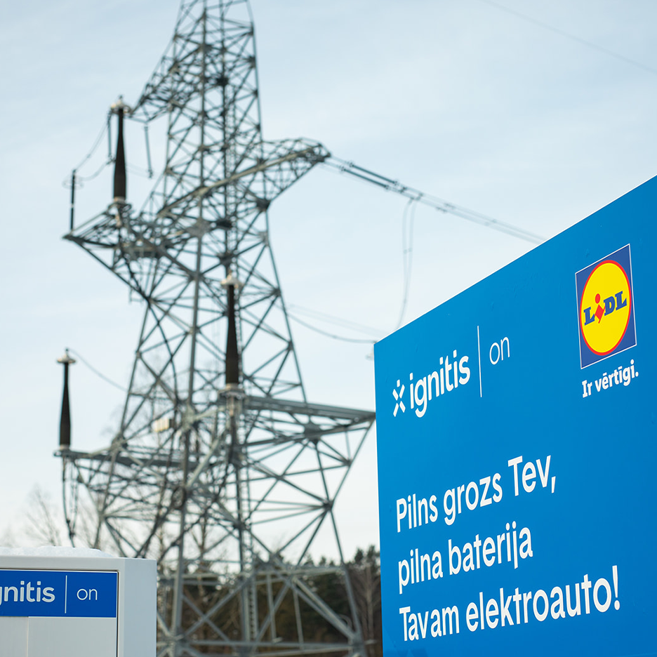 Reklāmas stends ar uzrakstu “Pilns grozs Tev, pilna baterija Tavam elektroauto!” uz elektropārvades līnijas fona.