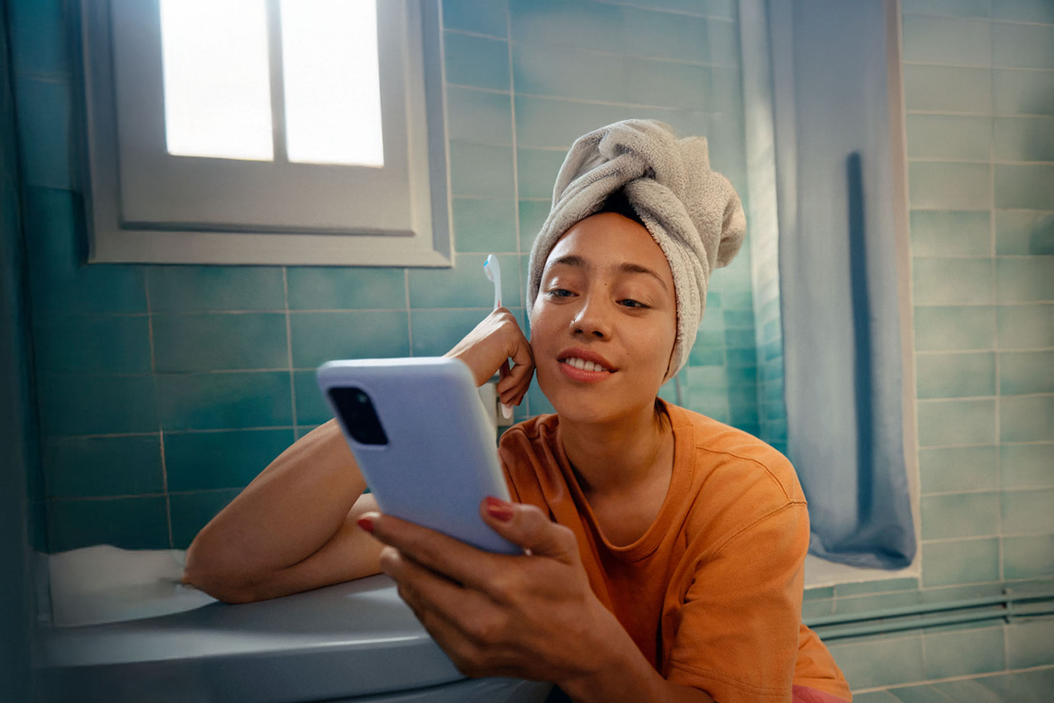 Sieviete ar dvieli uz galvas, izmantojot viedtālruni vannas istabā.