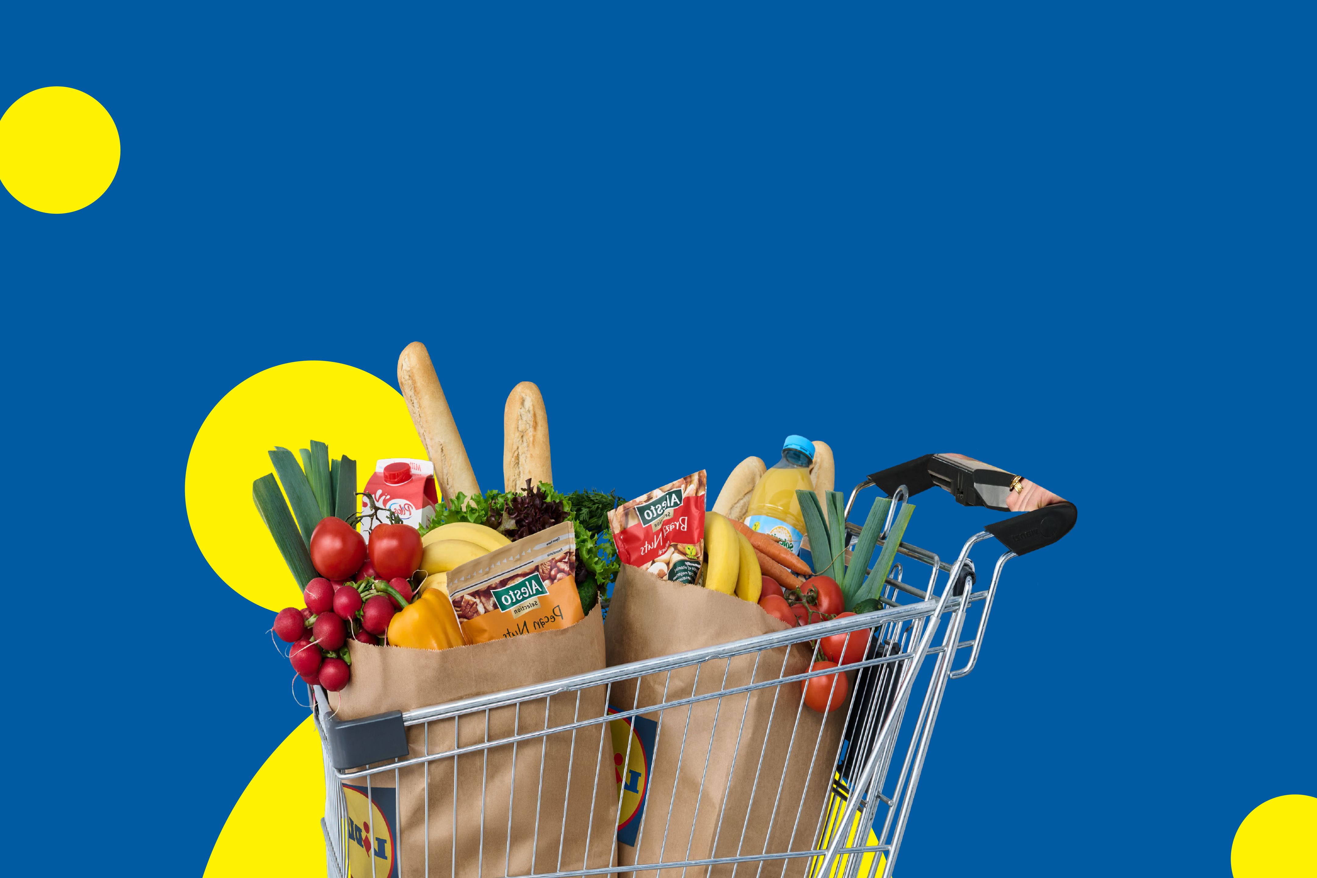 Iepirkumu ratiņi, kas piepildīti ar pārtikas precēm, piemēram, dārzeņiem, augļiem, maizi un riekstiem, uz zila fona ar dzelteniem apļiem.
