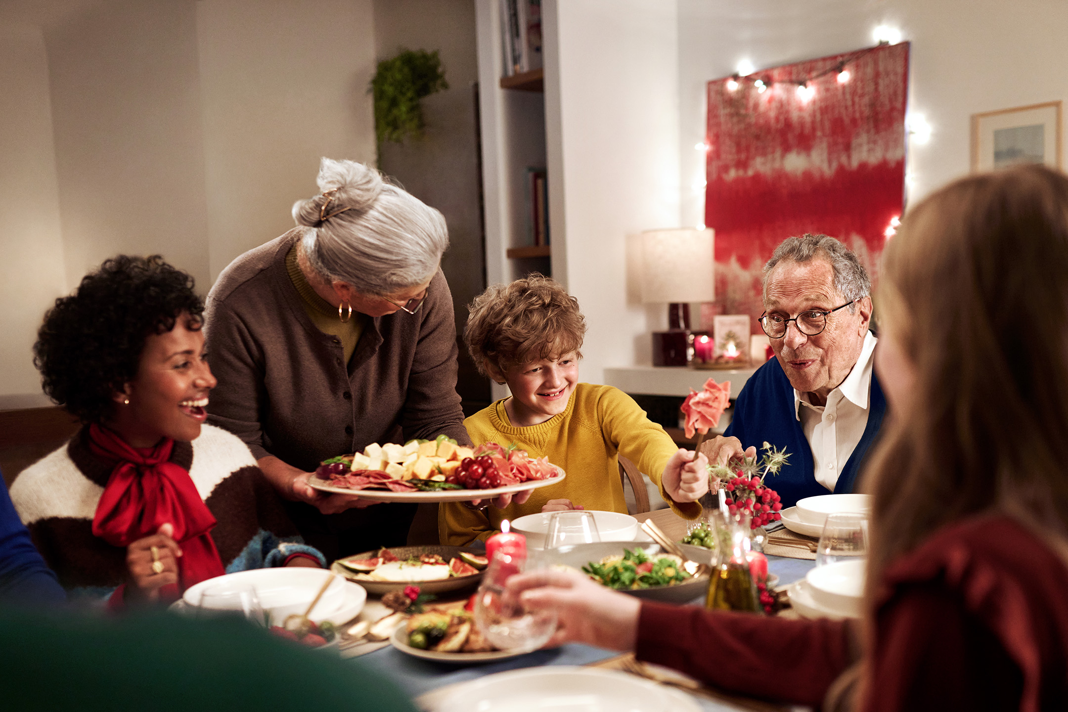 Ģimene pie svētku galda ar gaļas un siera uzkodām, salātiem un svecēm.