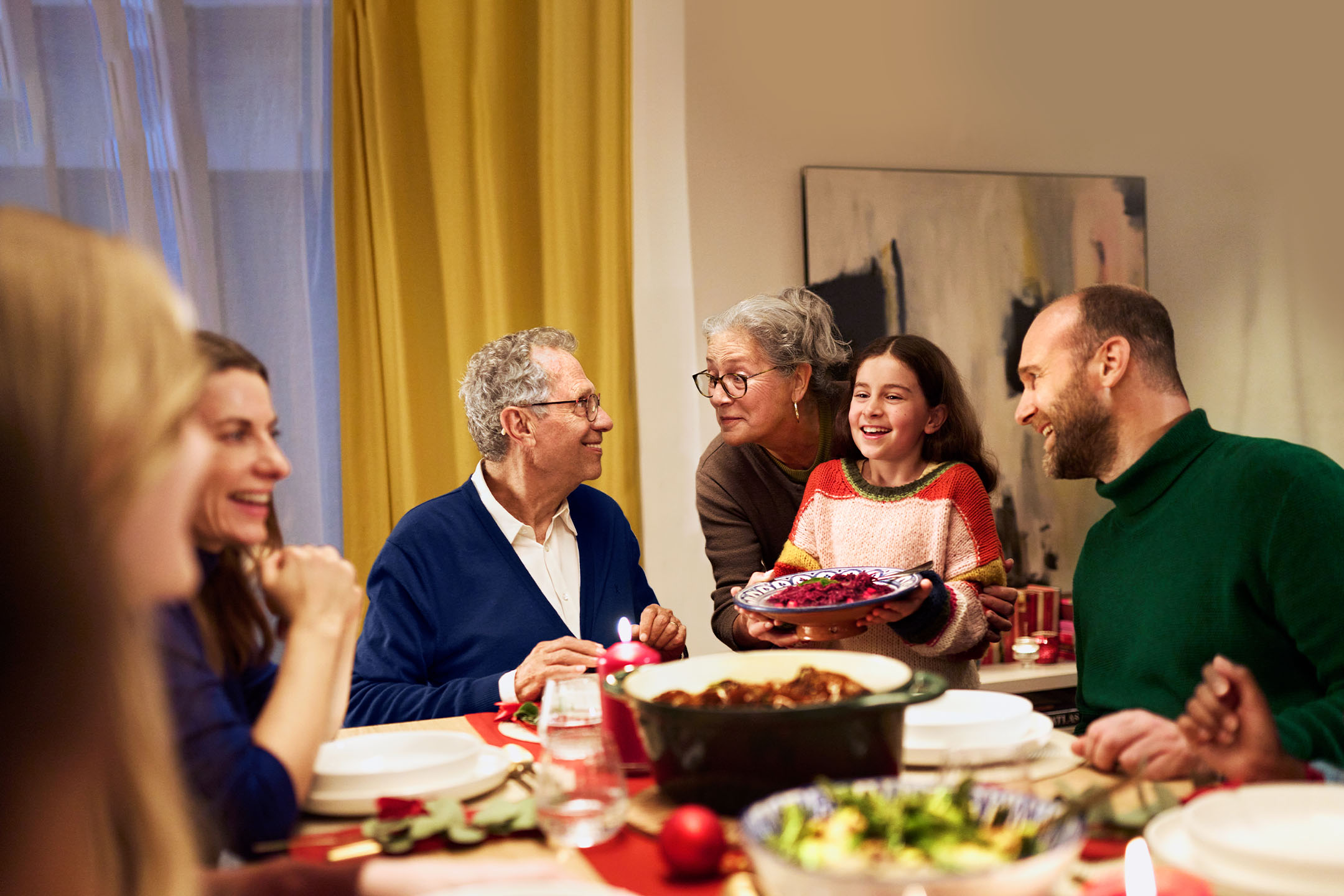 Ģimene pie galda, meitene pasniedz ēdienu, visi smaida un sarunājas.