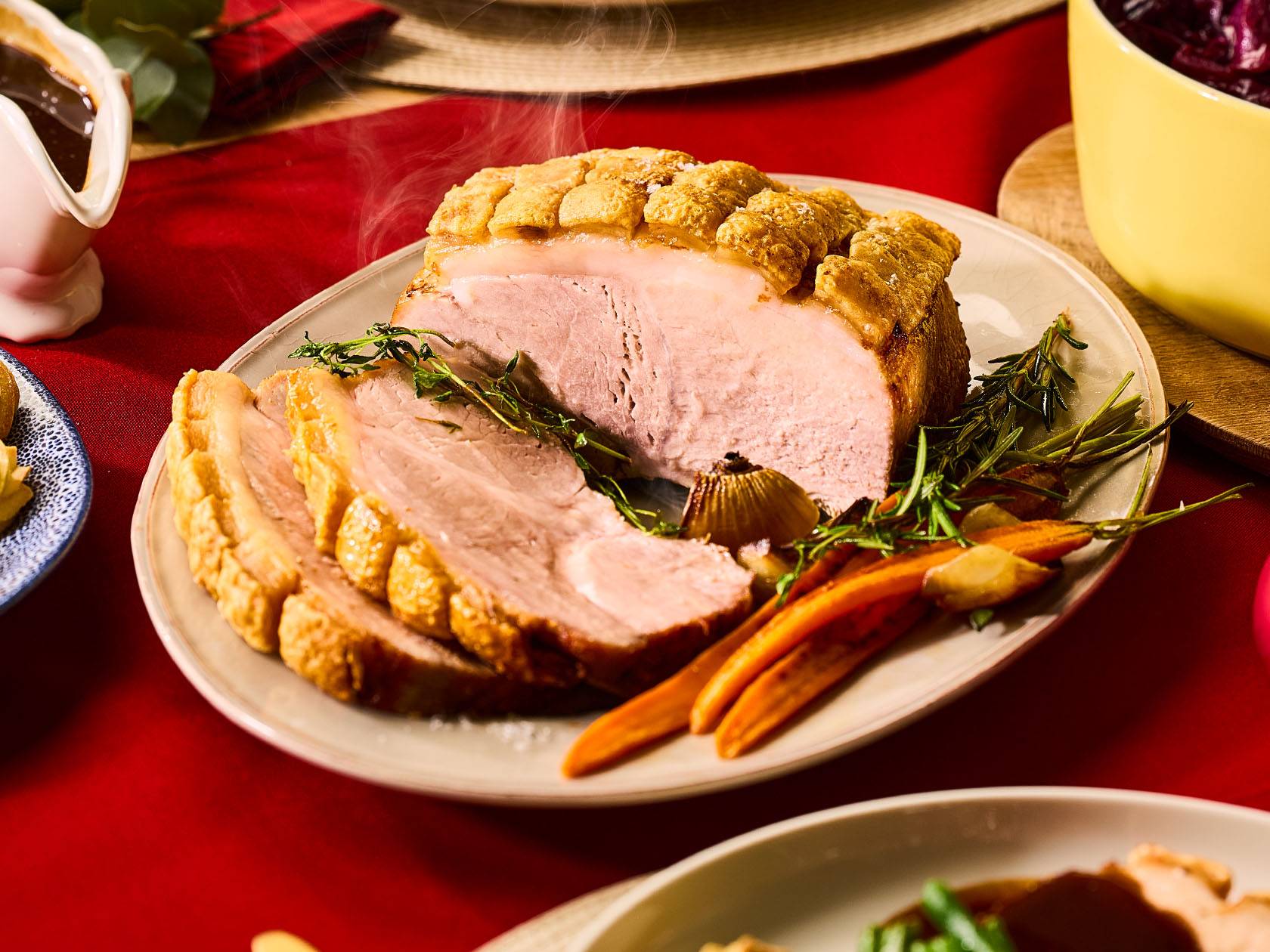 Cepta cūkgaļa ar kraukšķīgu garoziņu, sagriezta šķēlēs, ar dārzeņiem un mērci uz sarkana galdauta.